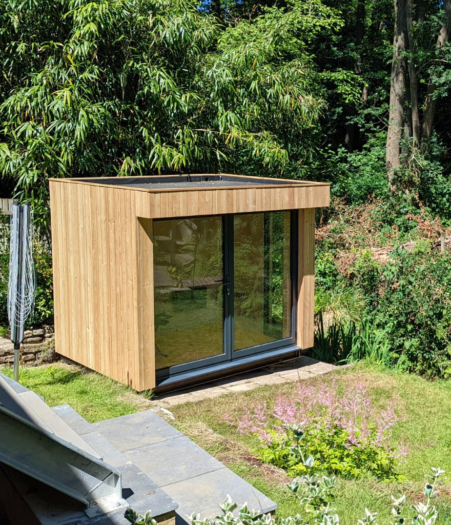 Compact garden office pod with vertical STK cedar cladding and large glass French doors, nestled in a lush green backyard with a small pink flower bed.