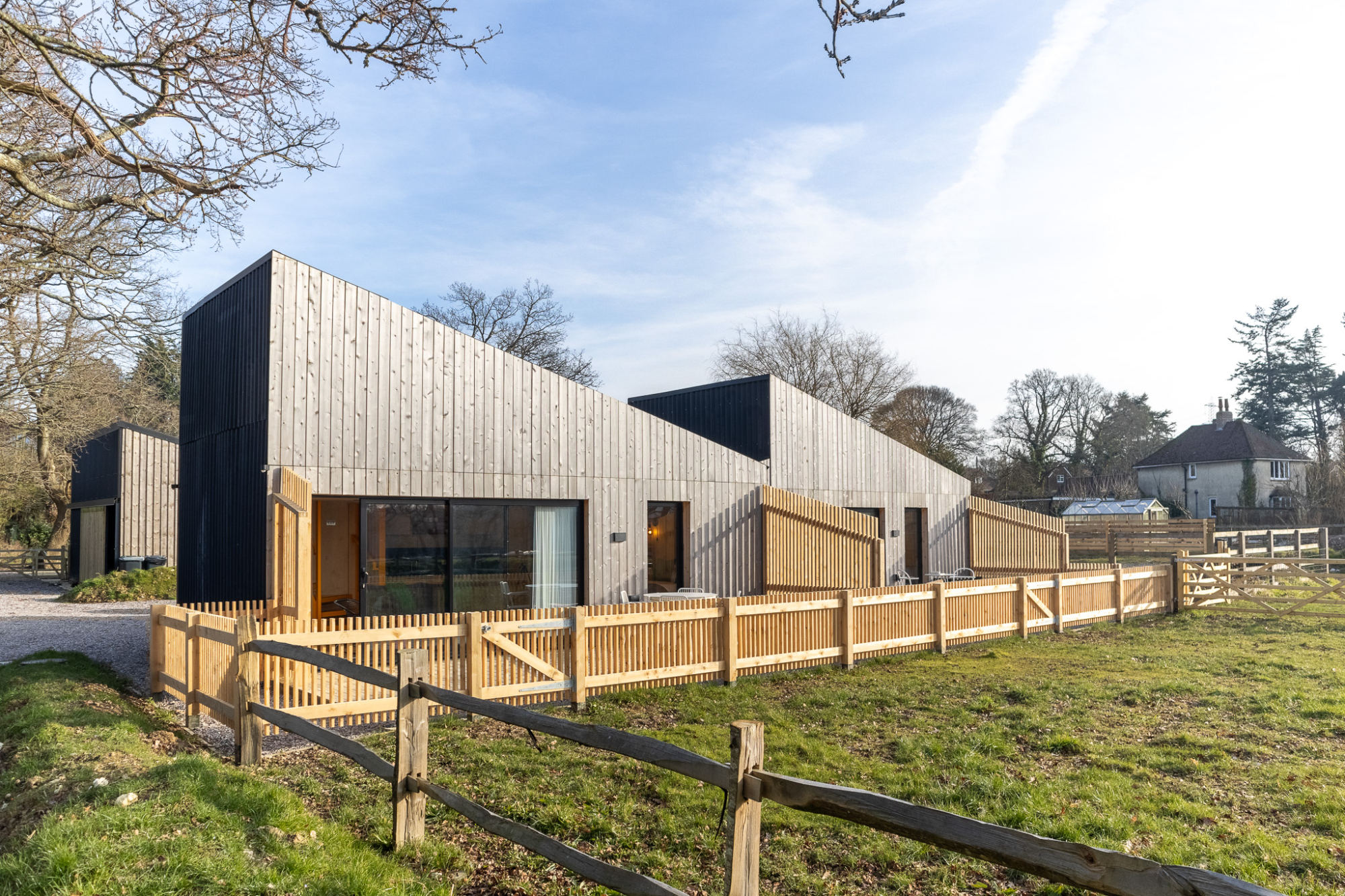 Modern timber holiday lodges with mono-pitch roofs and vertical wood cladding, featuring private fenced gardens overlooking a rural field under a blue sky.