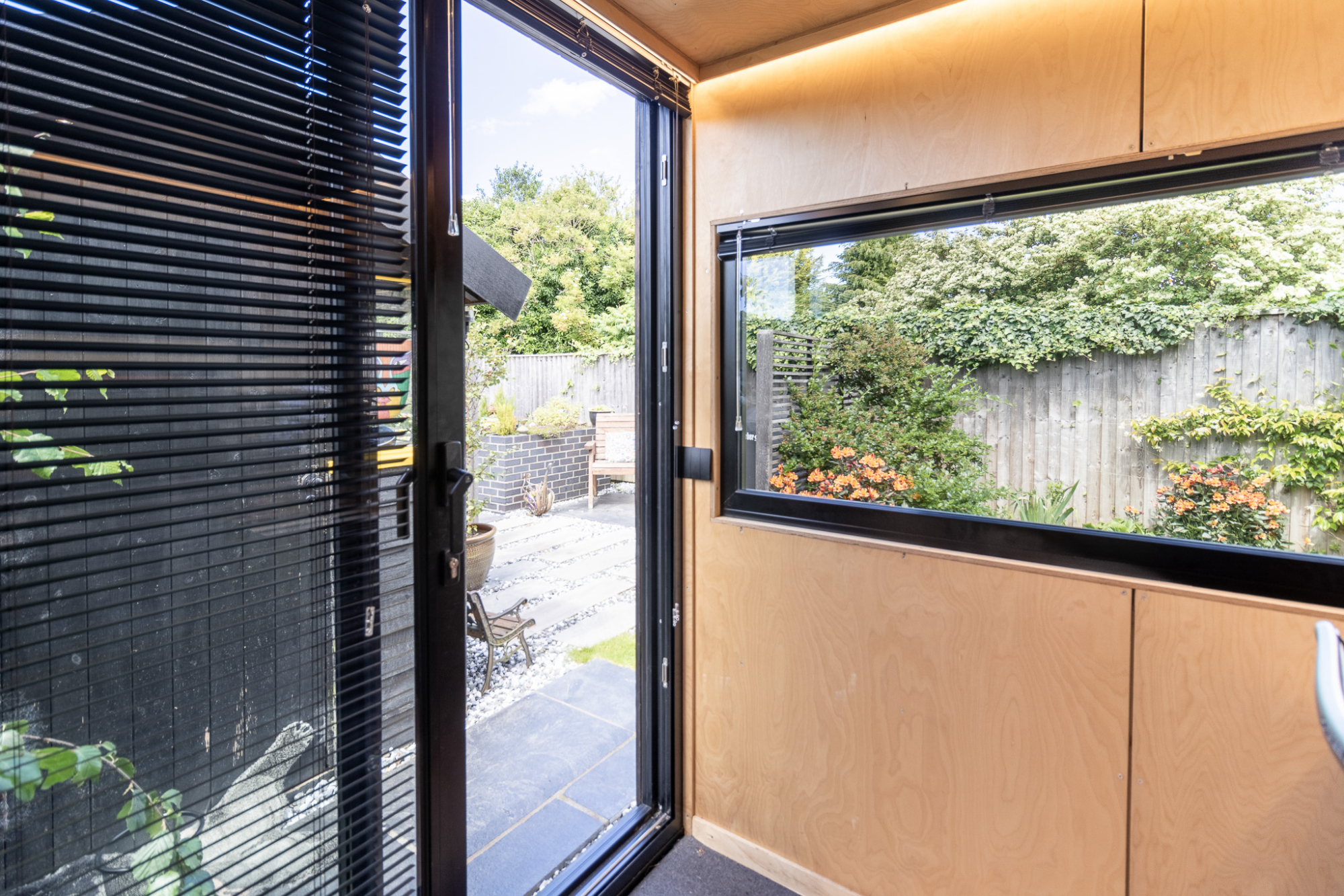 Interior of a modern garden office with plywood walls and a large picture window overlooking a landscaped backyard with a stone patio and wooden bench.