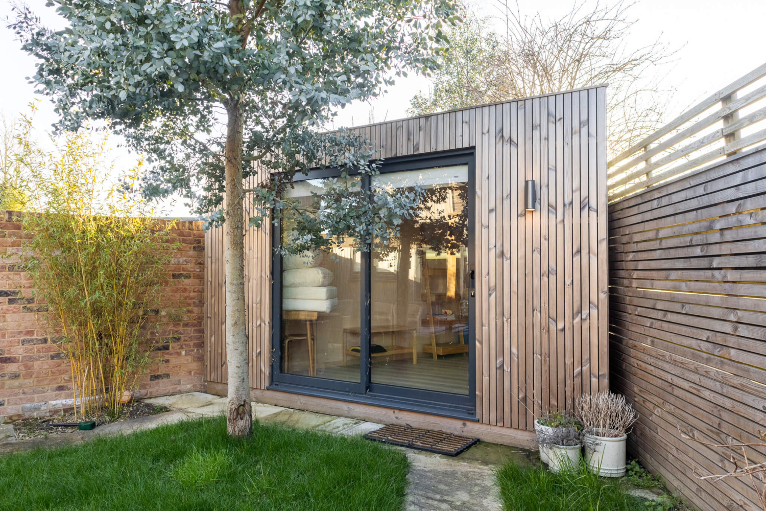 Modern plywood-lined garden office interior with a built-in desk, dual monitors, and views of a lush green backyard.