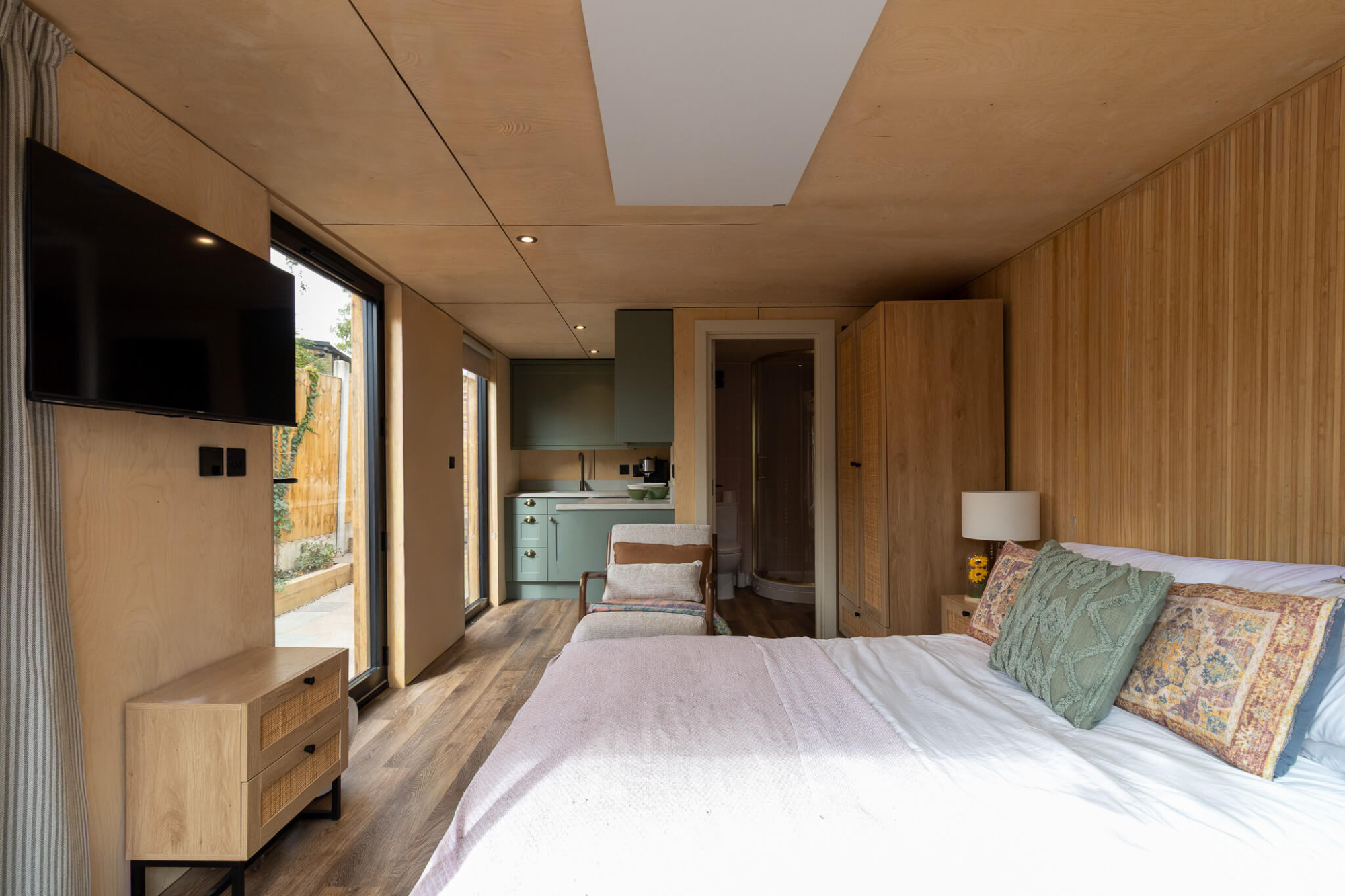 Interior of a luxury garden studio featuring a plywood-lined bedroom and living area, a modern sage green kitchenette, and a wall-mounted TV opposite a large glass door.