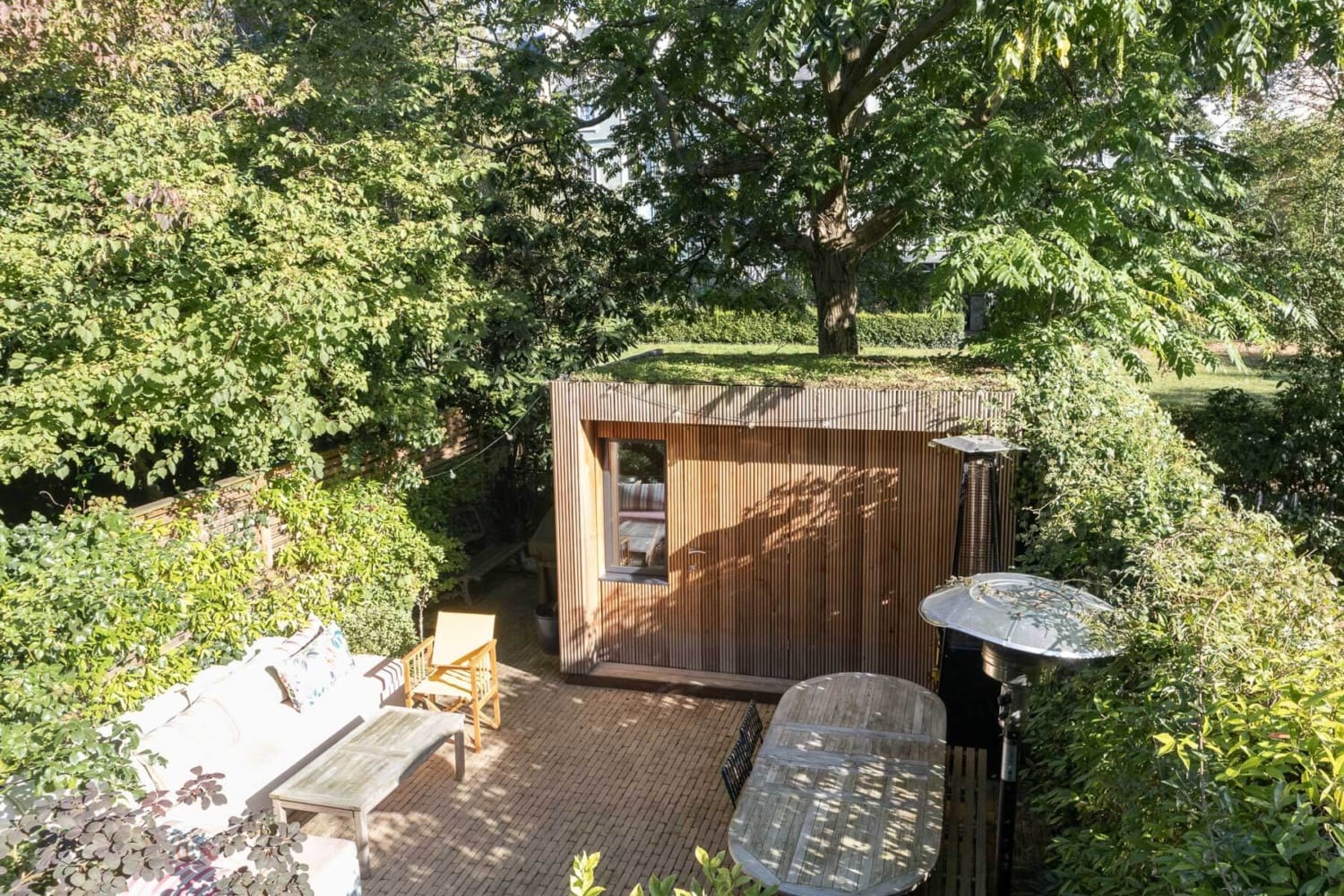 Modern wood-slatted garden sauna with a green sedum roof, surrounded by lush trees and a brick patio.