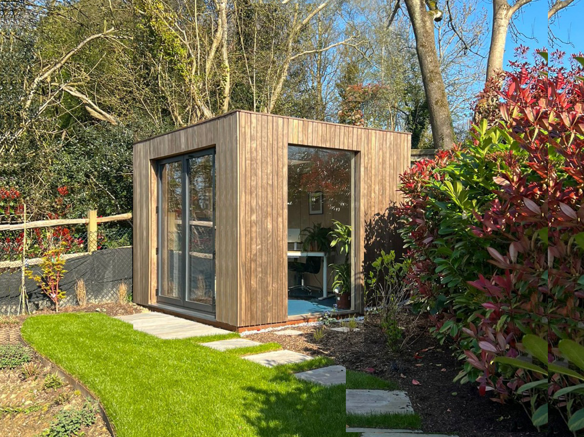 Spacious garden room interior with plywood walls, a large teal sectional sofa, a geometric rug, and black bifold glass doors.