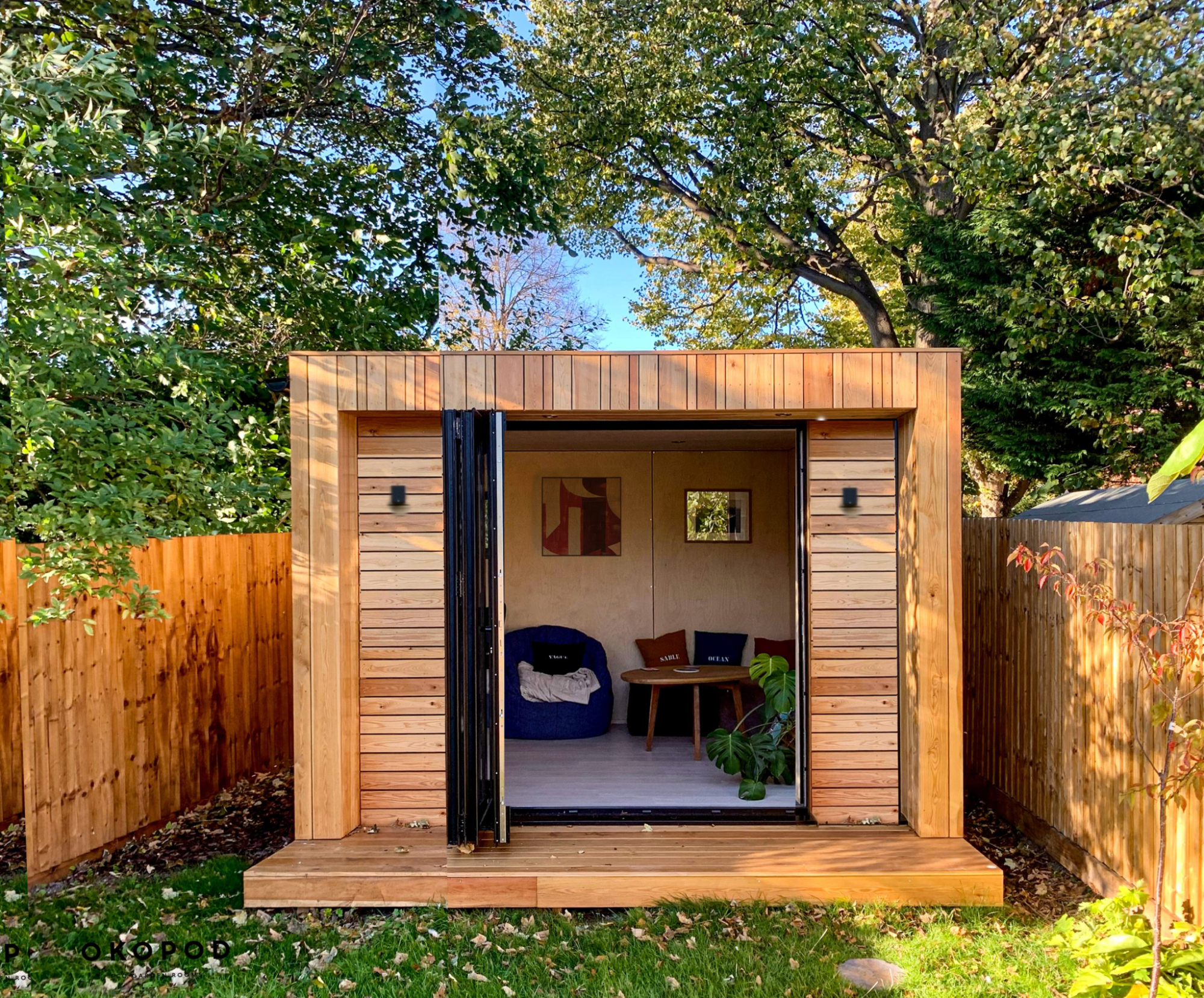 Compact cedar-clad garden pod with a small timber deck and black bifold doors, featuring a cozy interior with a blue bean bag and plywood walls.