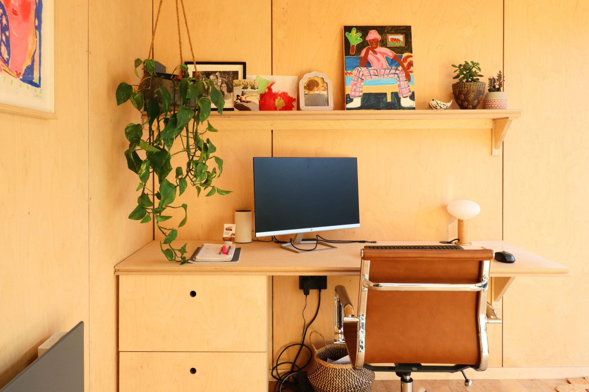 Interior of a modern plywood-lined garden office with a built-in desk, ergonomic chair, dual monitors, and views of a lush garden.