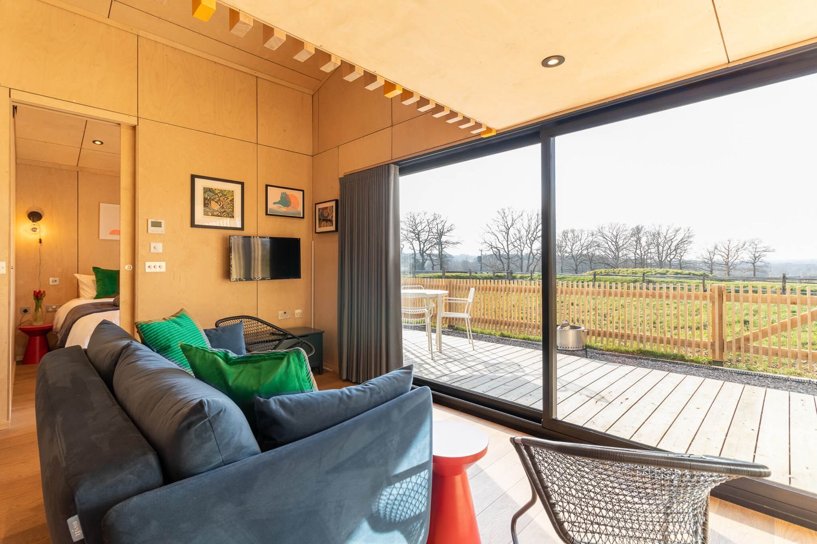 Cozy living room interior of a cabin, perfect for Airbnb or retreat accommodation on your land, featuring a blue sofa with green pillows, a wall-mounted TV, and large sliding glass doors opening to a wooden deck with outdoor seating and a scenic field view.