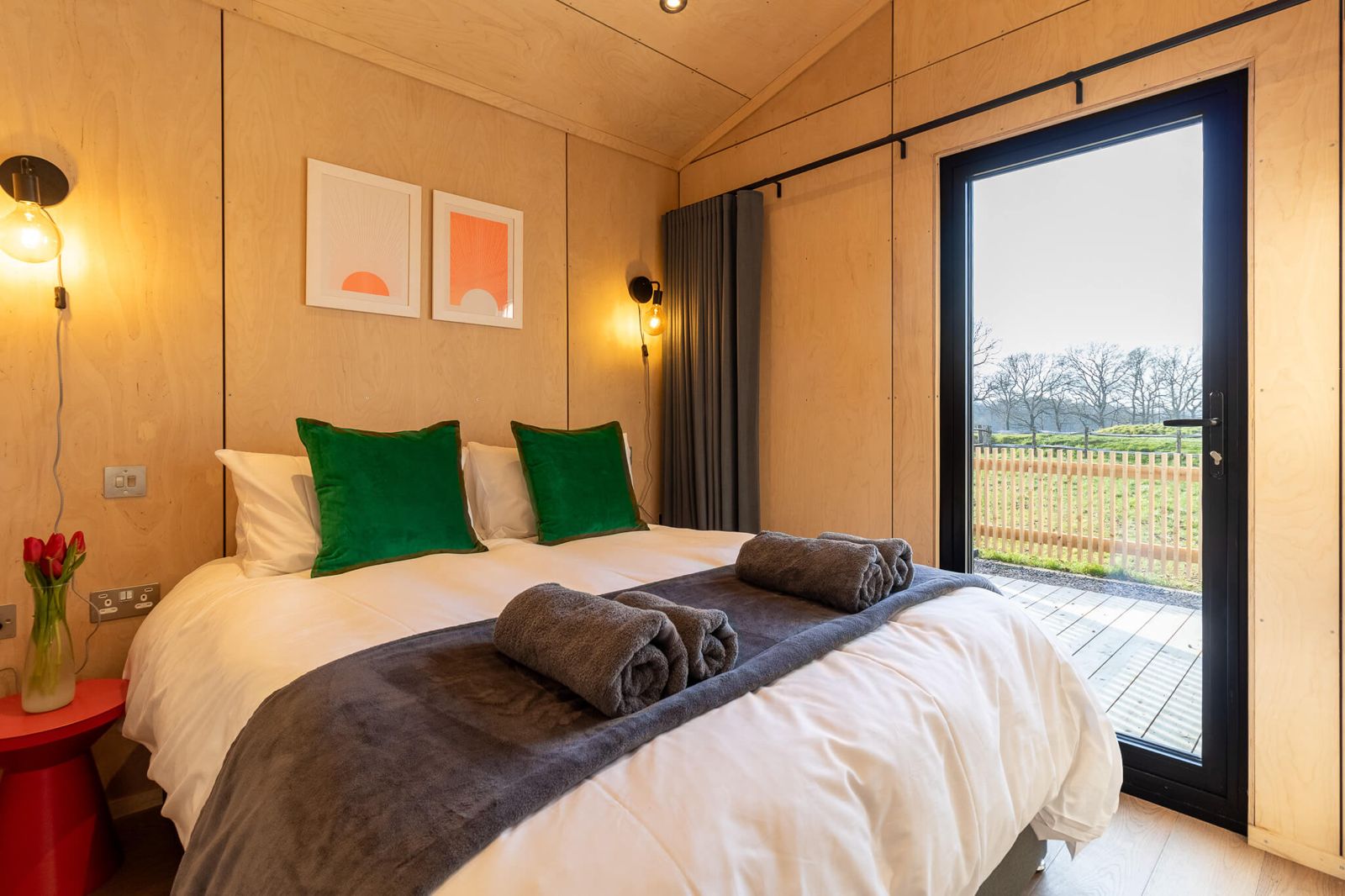 Cozy and bright bedroom in a cabin, perfectly suited for Airbnb or retreat accommodation on your land. The room features a comfortable bed with green pillows, natural wood walls, and a full-length glass door opening directly to an outdoor deck with a field view.