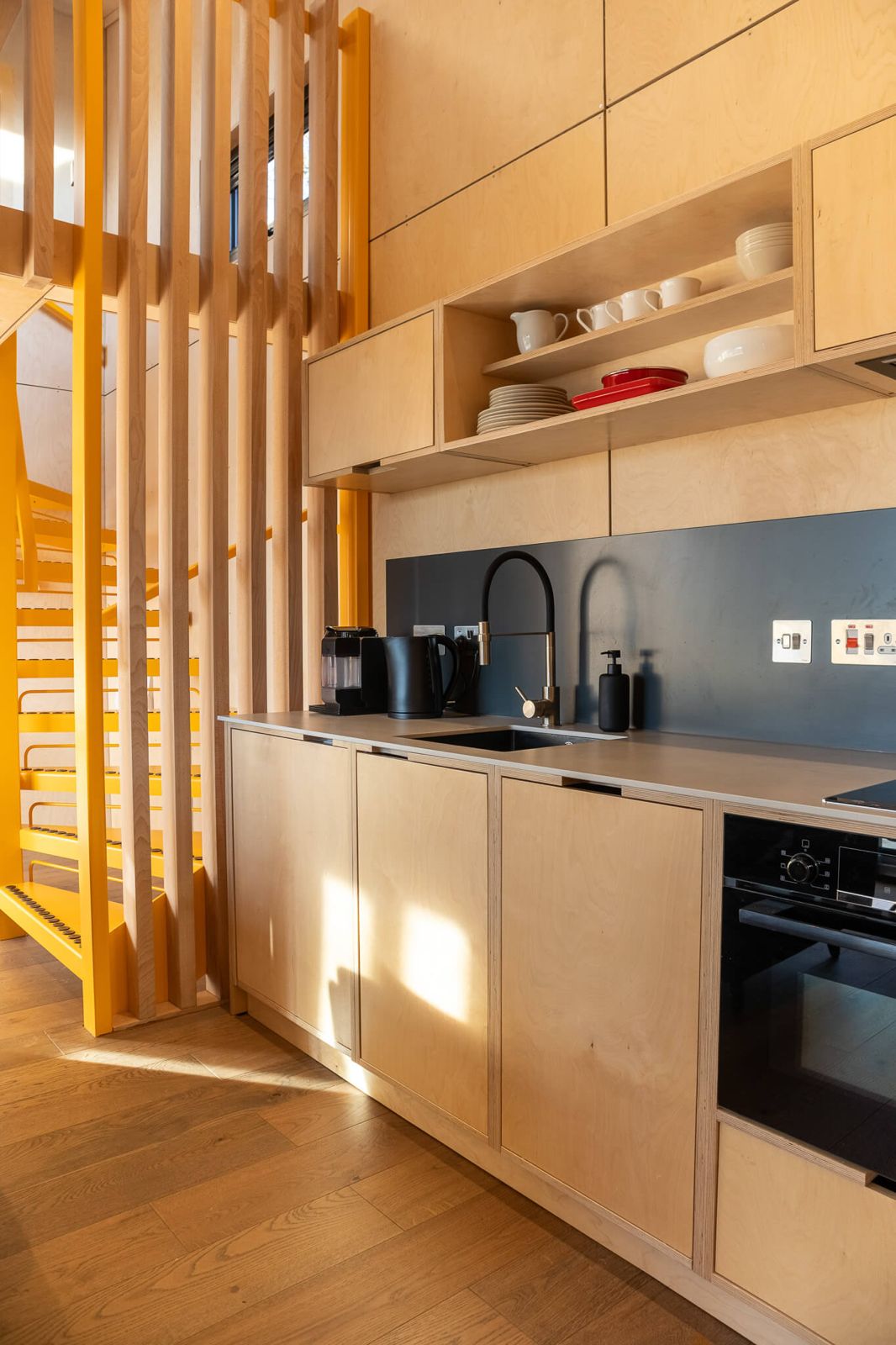 Compact and modern kitchen interior, ideal for Airbnb or retreat accommodation on your land, featuring light wood cabinetry, a black sink and faucet, and open shelving. A bright yellow spiral staircase and wooden slat partition are visible to the left.