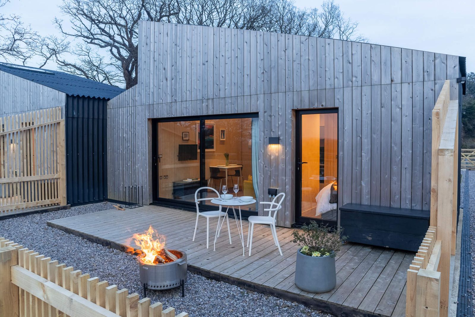 Exterior view of a modern timber-clad cabin with a private wooden deck and a fire pit, perfect for Airbnb or retreat accommodation on your land. Large glass doors and windows offer glimpses into the illuminated interior, hinting at comfortable living spaces.