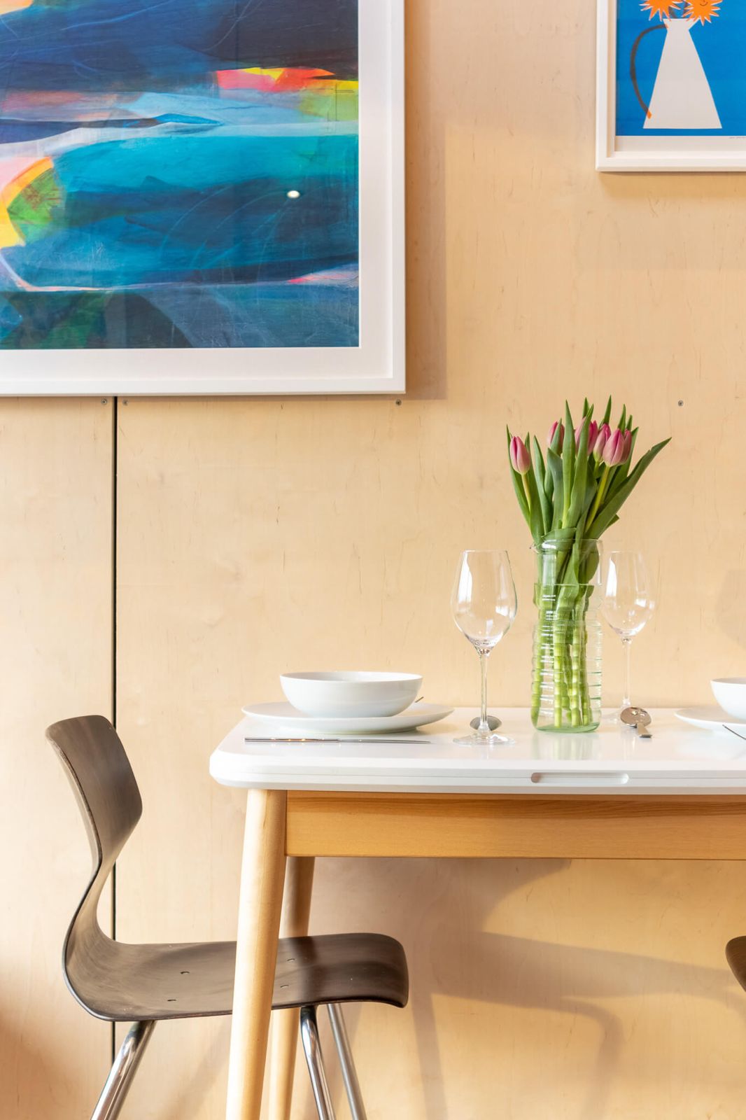 Cozy and stylish dining area within a cabin, featuring a white table set with glassware and a vase of pink tulips, ideal for Airbnb or retreat accommodation on your land. The space is adorned with colorful abstract art against natural wood-paneled walls.