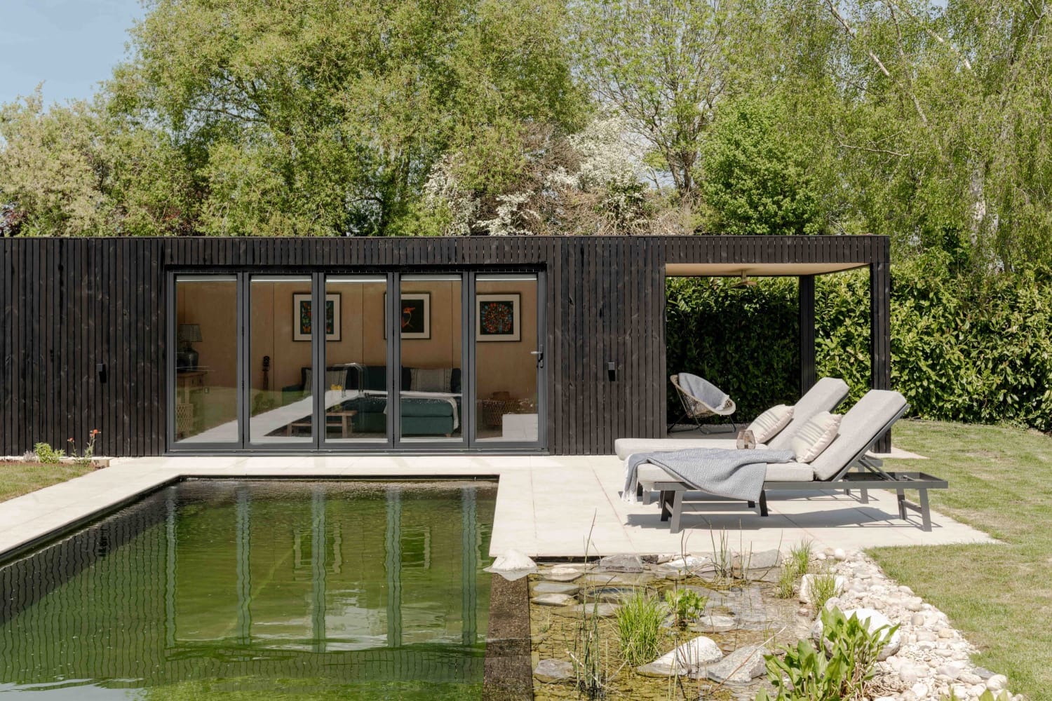 A minimalist Scandinavian garden pod featuring vertical light wood siding and floor-to-ceiling glass doors, integrated into a modern landscaped backyard with a stone patio.