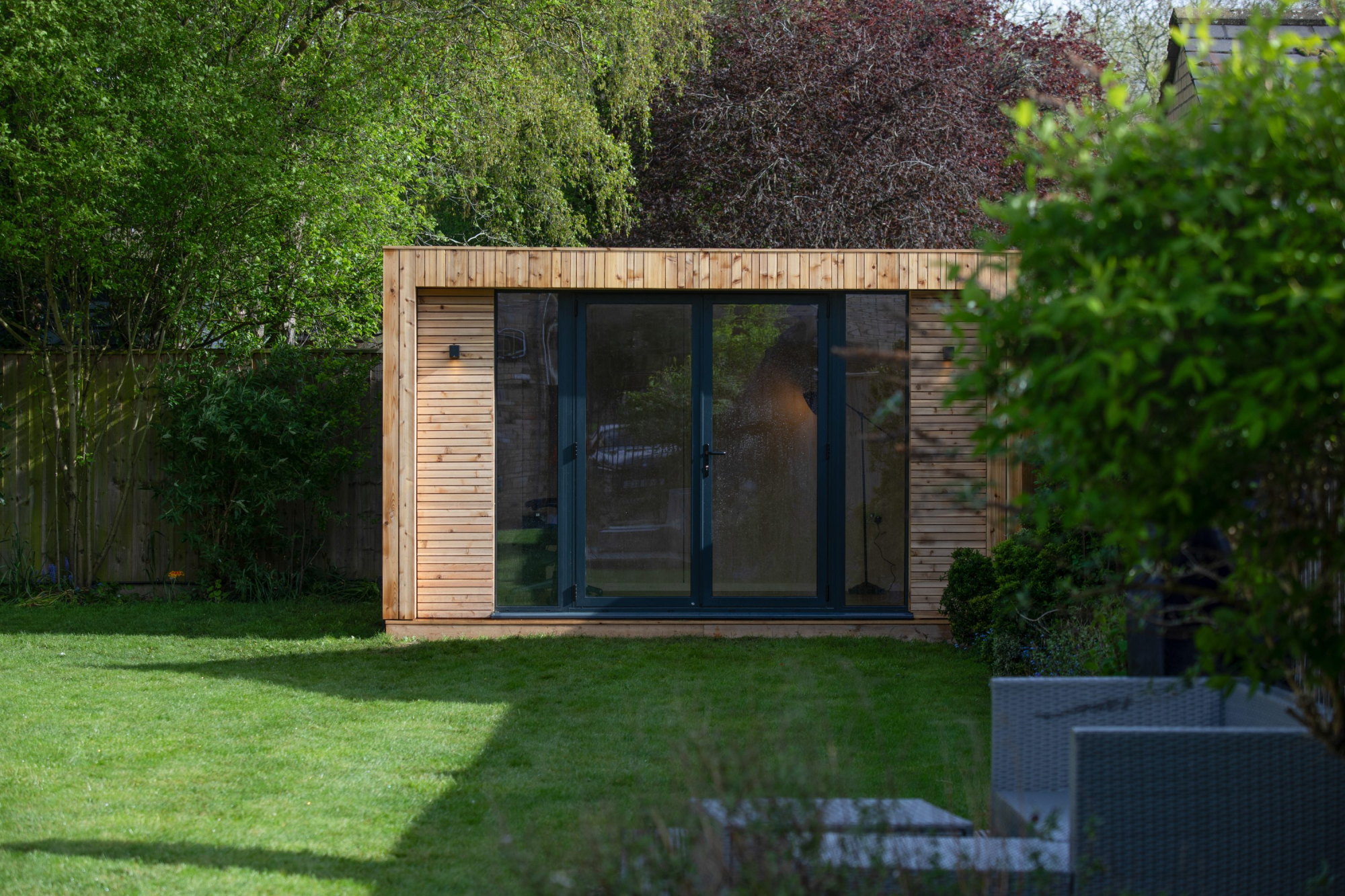 A beautiful cedar-clad small backyard office pod nestled on a manicured green lawn, featuring sleek glass double doors.