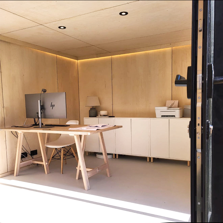 A minimalist interior design for a small backyard office featuring a wooden trestle desk, warm accent lighting, and clean white cabinets.