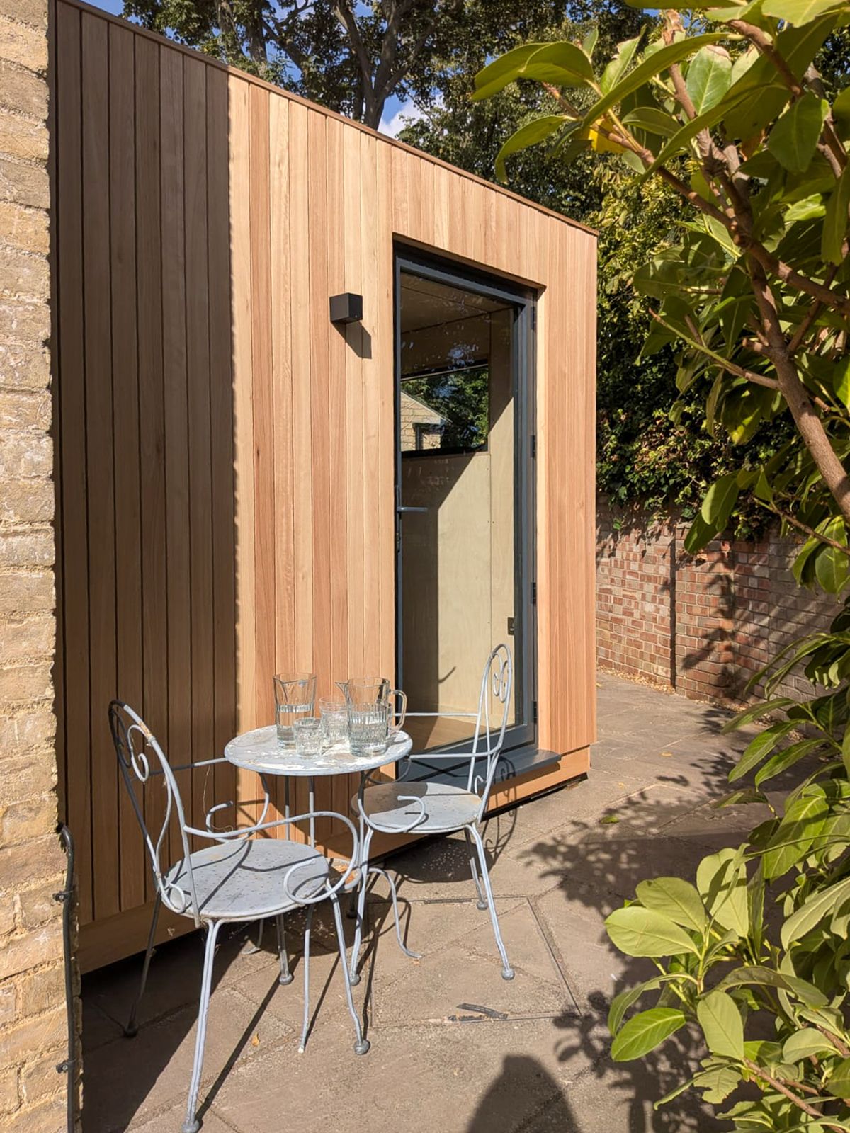 Close-up side view of a prefabricated classroom pod showing vertical wood paneling and entrance next to outdoor patio furniture.