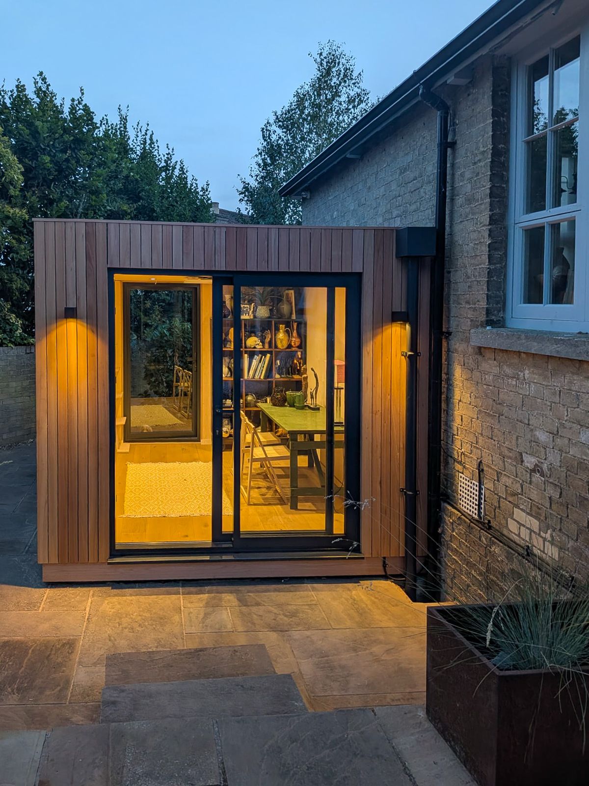 Exterior view at dusk of an illuminated timber-clad prefabricated classroom pod with warm interior lighting and glass sliding doors.